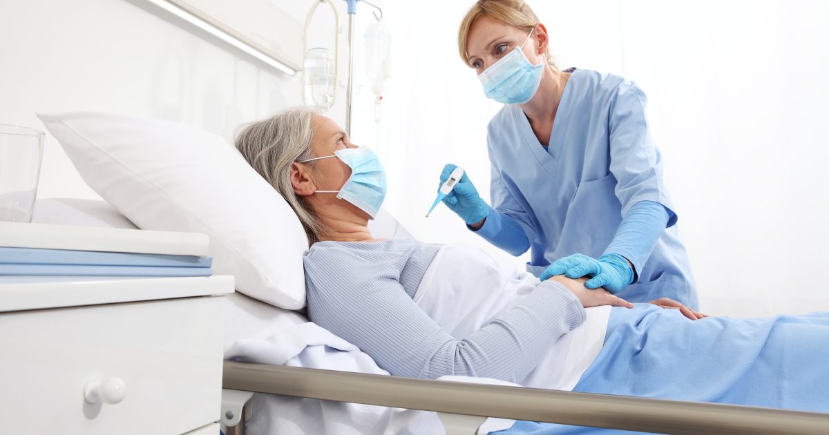 nurse with thermometer measures fever on elderly woman patient lying in the hospital room bed, wearing protective gloves and medical surgical mask, coronavirus covid 19 protection concept