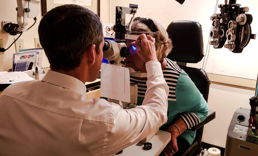 Image of an eye exam room with a woman sitting in a chair, a doctor standing next to her, and various eye exam equipment in the background.