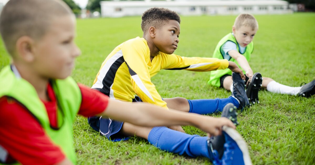 Three children in colorful soccer uniforms stretch their legs on a grassy field before practice