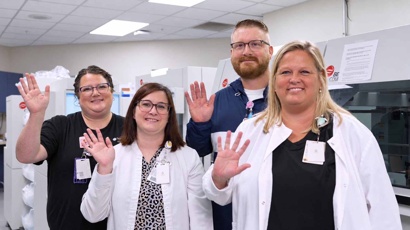 Four smiling laboratory staff members wave at the camera while standing in front of lab equipment.